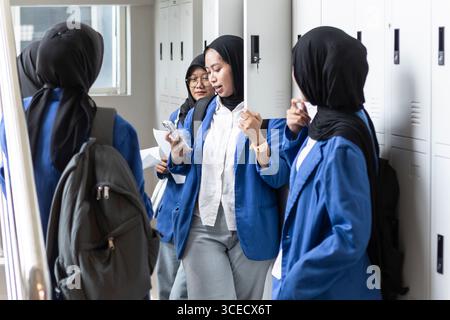 A group of Asian Muslim students in blue uniforms stand near lockers as they return to school. They are engaged with each other, using their phones an Stock Photo