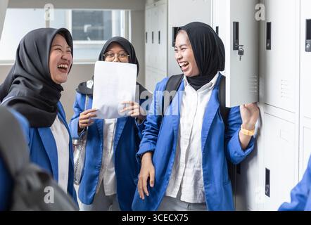 A group of Asian Muslim students in a school corridor, laughing and sharing a moment of camaraderie. They wear blue uniforms and carry backpacks, read Stock Photo