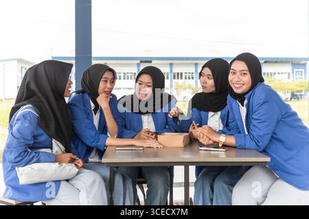 A group of Asian Muslim students in headscarves sit together at an outdoor table, engaged in a collaborative study session, emphasizing education and Stock Photo
