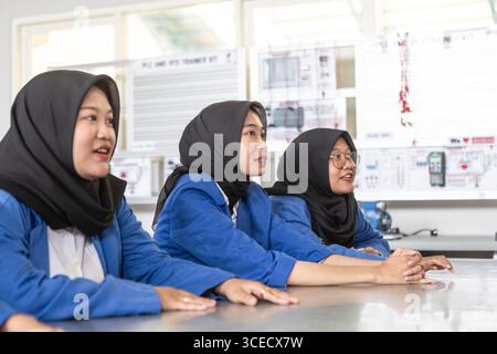Three Asian Muslim students in hijabs and blue uniforms sit attentively at a classroom desk, immersing in a learning environment, ready for new academ Stock Photo