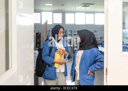 Two Asian Muslim students in blue uniforms, conversing as they enter a bright classroom. They seem engaged and ready to continue their education journ Stock Photo