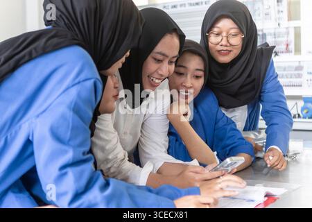 Asian Muslim teacher engaging with students using a smartphone in a classroom setting. The students, wearing blue uniforms, gather around the teacher, Stock Photo
