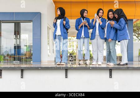 Asian Muslim students in hijabs and blue blazers gather at an educational institution, engaging in conversation and using smartphones as they prepare Stock Photo