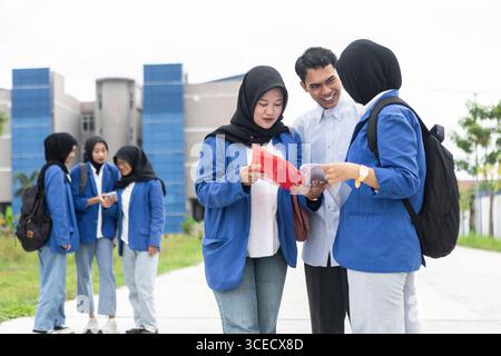A group of Asian Muslim students in blue jackets and hijabs stand together, engaging in a lively discussion as they return to school. They hold books, Stock Photo