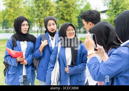 A group of Asian Muslim students, wearing hijabs and matching blue uniforms, gather outdoors with backpacks and papers, happily chatting and interacti Stock Photo