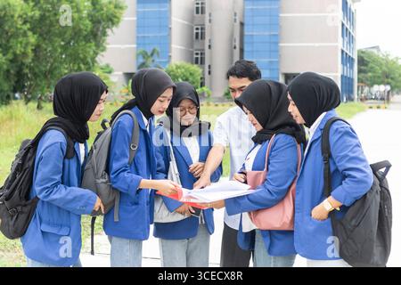 A group of Asian Muslim students in blue uniforms gathers outside a modern school building, engaged in a collaborative learning activity, symbolizing Stock Photo