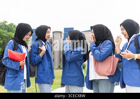 A group of Asian Muslim students wearing hijabs and blue uniforms, enjoying a lively conversation while standing together outdoors, as they return to Stock Photo