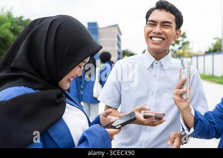 A group of Asian students, including a Muslim woman in hijab, enjoy a lively conversation and laughter on a vibrant school campus, celebrating friends Stock Photo
