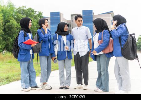 A group of Asian Muslim students in casual attire, wearing backpacks and carrying materials, are gathering outside a school building, symbolizing a re Stock Photo