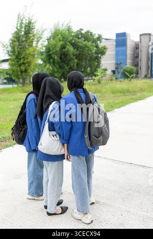Three Asian Muslim women in blue uniforms stand together with backpacks, preparing for a new academic year. The outdoor setting highlights their schoo Stock Photo