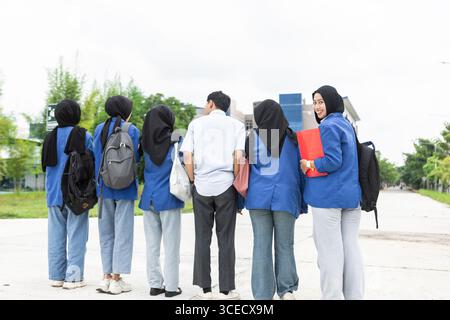 A group of Asian Muslim students, wearing blue uniforms and backpacks, happily head back to school. They walk along a campus pathway, ready to embrace Stock Photo