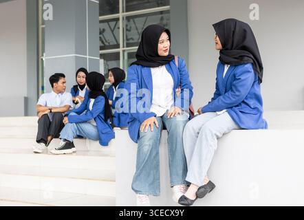 Asian Muslim students wearing hijabs and casual attire engage in conversation while sitting together in front of a modern school building, expressing Stock Photo