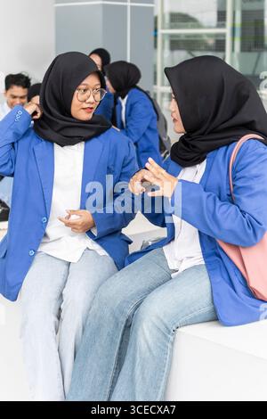 Two Asian Muslim students, wearing black hijabs and blue jackets, engage in conversation while seated on a bench. They appear to be students back at c Stock Photo