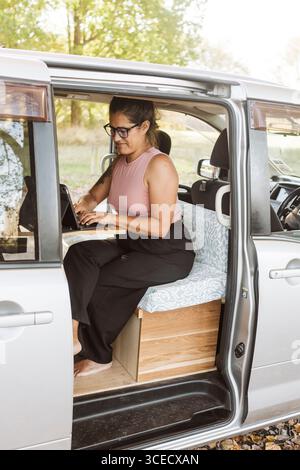 A woman works remotely from a cozy van in the scenic autumn setting of New Zealand. She is focused on her laptop, surrounded by colorful foliage and n Stock Photo