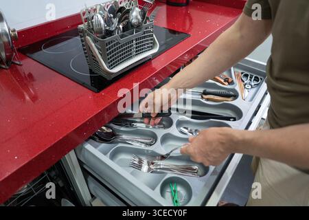 A person organizes utensils in a kitchen drawer, sorting knives, forks, and spoons in a modern home with a red countertop, emphasizing household order Stock Photo