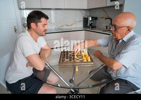A father and adult son engage in a friendly game of chess at home, strengthening their bond. The intimate setting highlights quality intergenerational Stock Photo