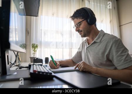 A man sits at his desk wearing headphones, focused on learning Mandarin online. He takes notes in a notepad with a pen, engaging in language studies f Stock Photo