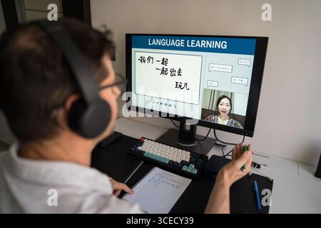 A man is engaged in an online Mandarin language lesson at home, wearing headphones and taking notes. The computer screen displays Chinese characters a Stock Photo