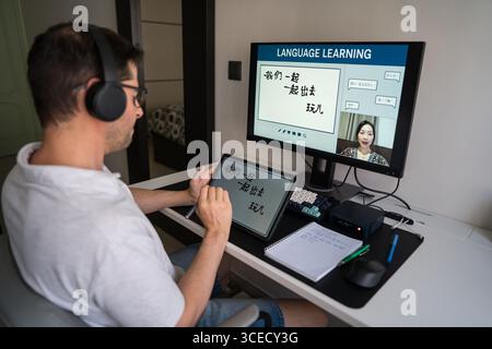 A man wearing headphones studies Mandarin online at home, using a digital tablet and computer setup. The screen displays a virtual language lesson wit Stock Photo