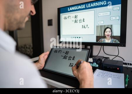 A man learning Mandarin online at home, engaging with a virtual instructor via video call. He writes Chinese characters on a digital tablet, immersed Stock Photo
