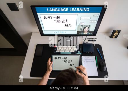 A man is engaged in learning Mandarin through an online course at home, using a computer and tablet setup for interactive lessons with a virtual teach Stock Photo