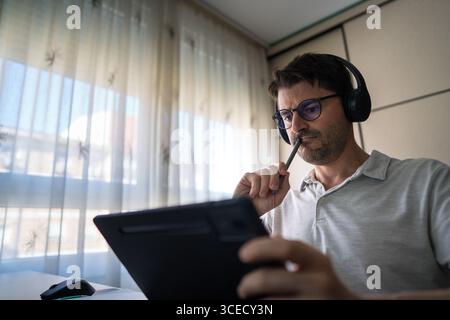 A man sits at home, equipped with headphones, focusing intently on studying Mandarin using a digital tablet, illustrating the modern approach to langu Stock Photo