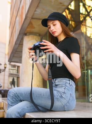 A woman photographer sits on a city bench, focused on her camera. Dressed in casual attire with a black cap, her thoughtful expression highlights her Stock Photo