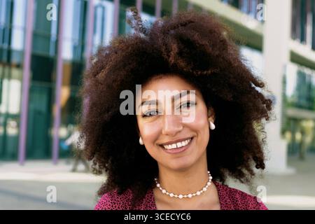 A young Arab woman with a bright smile and voluminous afro hair stands outdoors looking at camera, wearing a pink patterned top and pearl jewelry. The Stock Photo