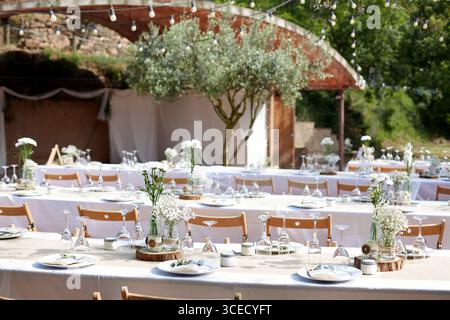 Elegant boho wedding table setup with white flowers in glass vases on wooden tables and chairs. String lights and greenery create a romantic and pictu Stock Photo