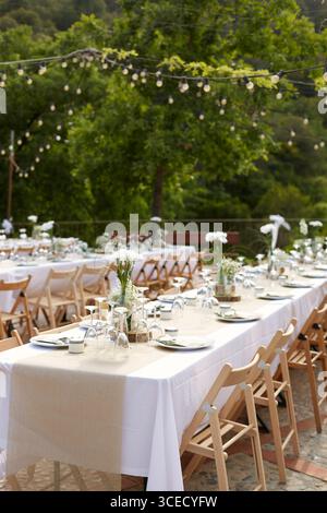 An elegantly set outdoor wedding table features white tablecloths and rustic burlap runners. Surrounded by natural greenery, it is adorned with simple Stock Photo