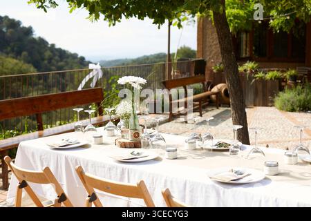 Elegant outdoor wedding table adorned with white floral decorations in glass vases on a wooden table. Overlooking a scenic view, the setup is both rom Stock Photo
