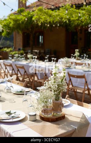 Charming outdoor wedding table adorned with white floral centerpieces in rustic jars, set on natural linen table runners. Elegant glassware and wooden Stock Photo