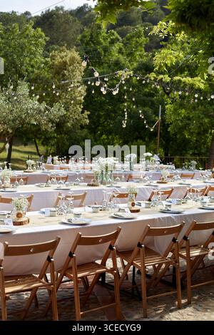 Elegant outdoor wedding setup with wooden chairs and tables, adorned with white tablecloths and glass vases holding delicate floral arrangements, unde Stock Photo