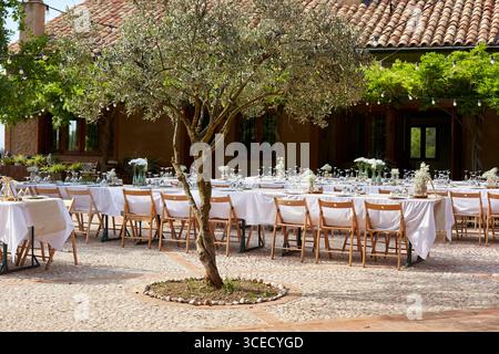 Charming outdoor wedding reception with long tables set with white linens, surrounded by wooden chairs. Rustic charm enhanced by an old tree, cobblest Stock Photo