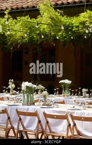 An elegant outdoor wedding setting features long tables adorned with white tablecloths, wooden chairs, and delicate floral arrangements with white flo Stock Photo