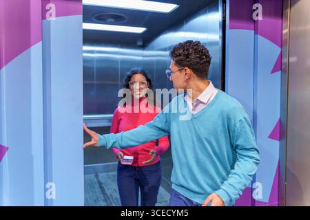 A Gen Z man holds the elevator door open for a happy woman holding a smartphone. They are casually dressed and smiling, showcasing modern social etiqu Stock Photo