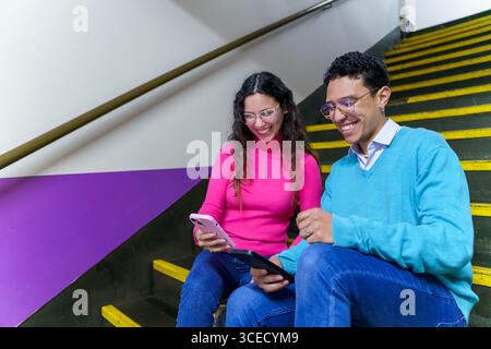 A cheerful smiling young woman and man, both Gen Z, smile while using a smartphone and tablet on a staircase, showcasing modern digital communication Stock Photo