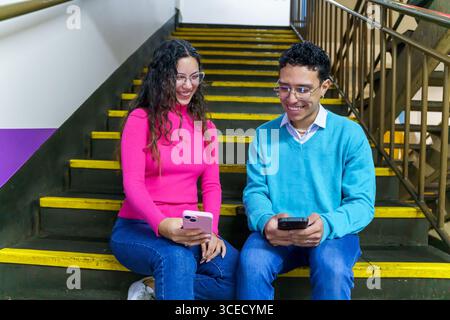 A cheerful young woman and man from Gen Z sit on stairs, smiling as they engage with smartphones. Their interaction highlights a modern digital lifest Stock Photo