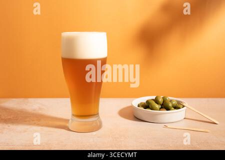 A tall glass of frothy lager beer sits on a light-colored surface, next to a bowl of green olives with a wooden pick. The backdrop is a warm orange hu Stock Photo