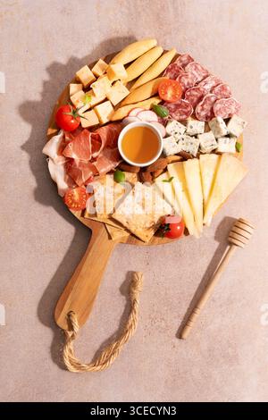 Top view of a cheese platter on wooden board featuring assorted cheeses, cured meats, tomatoes, nuts, and honey dip, set against a rustic stone backgr Stock Photo