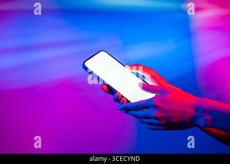 A hand holding a smartphone, illuminated by colorful neon lights. The glowing screen contrasts vividly with the purple and blue hues, creating a moder Stock Photo