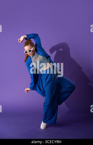 A young woman dancer striking a dynamic pose against a vivid purple background. Her contemporary blue outfit contrasts with her elegant dance stance, Stock Photo