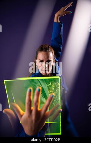 A young woman engages with a neon green futuristic device, highlighting modern technological innovations and concepts, set against a vibrant purple ba Stock Photo