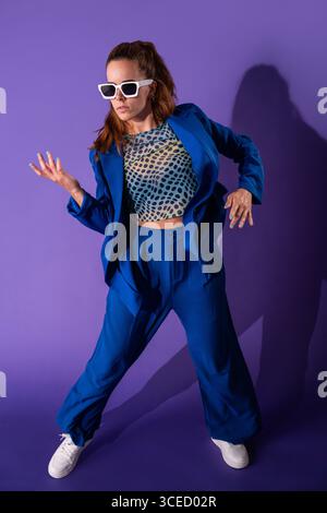 A young woman dancer striking a dynamic pose against a vibrant purple background. She displays energy and style, embodying the essence of modern dance Stock Photo