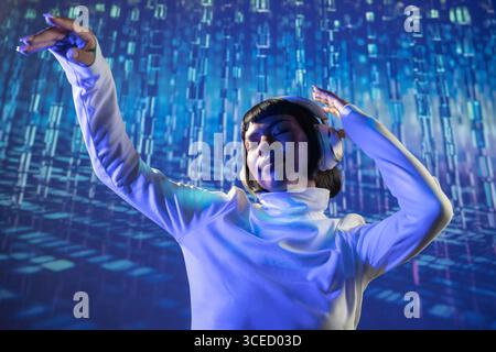 A futuristic Latin woman immersed in music, wearing headphones and dressed in a white turtleneck, surrounded by a digital backdrop of blue and purple Stock Photo
