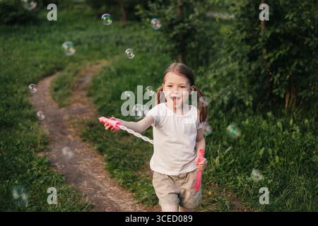 A joyful girl embraces summer play, running through a lush green garden while blowing and chasing sparkling soap bubbles, capturing the essence of chi Stock Photo