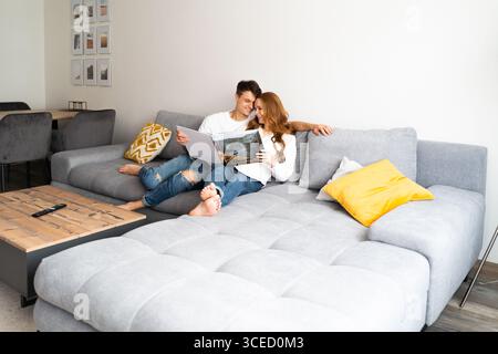 A joyful young couple relaxes on a cozy gray sofa, engaged in reading a large photo album. The warm ambiance highlights their excitement and anticipat Stock Photo