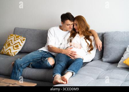 A young couple sits closely on a comfortable couch, with the man gently touching the woman's pregnant belly. They appear relaxed and happy, embracing Stock Photo