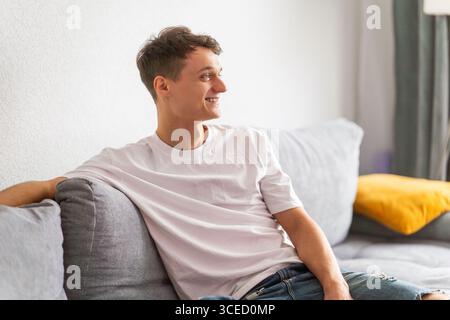 A man wearing a white t-shirt sits comfortably on a grey sofa, smiling and looking away, enjoying a relaxing moment at home with a yellow cushion in t Stock Photo