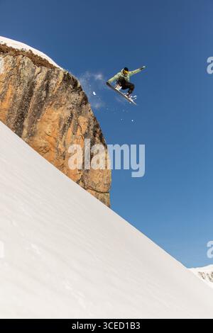 A daring snowboarder executes an exhilarating jump off a snow-covered cliff, soaring high against a clear blue sky in a stunning winter mountain lands Stock Photo